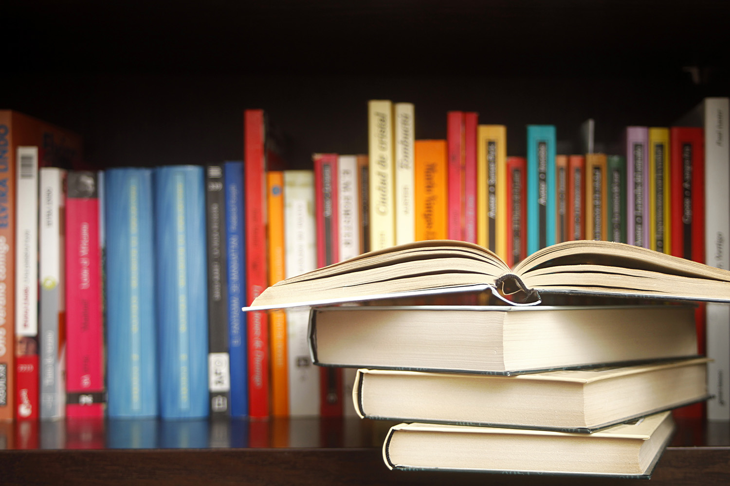 Stack of books on a wooden library shelf, the one on top open, multicolored book spines in a row in the background