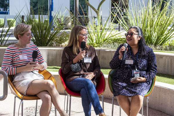 Three women sit in chairs in a small amphitheater and speak into microphones