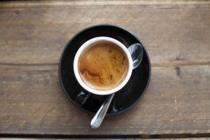 A small cup of espresso sits on a black saucer with a teaspoon on a wooden tabletop