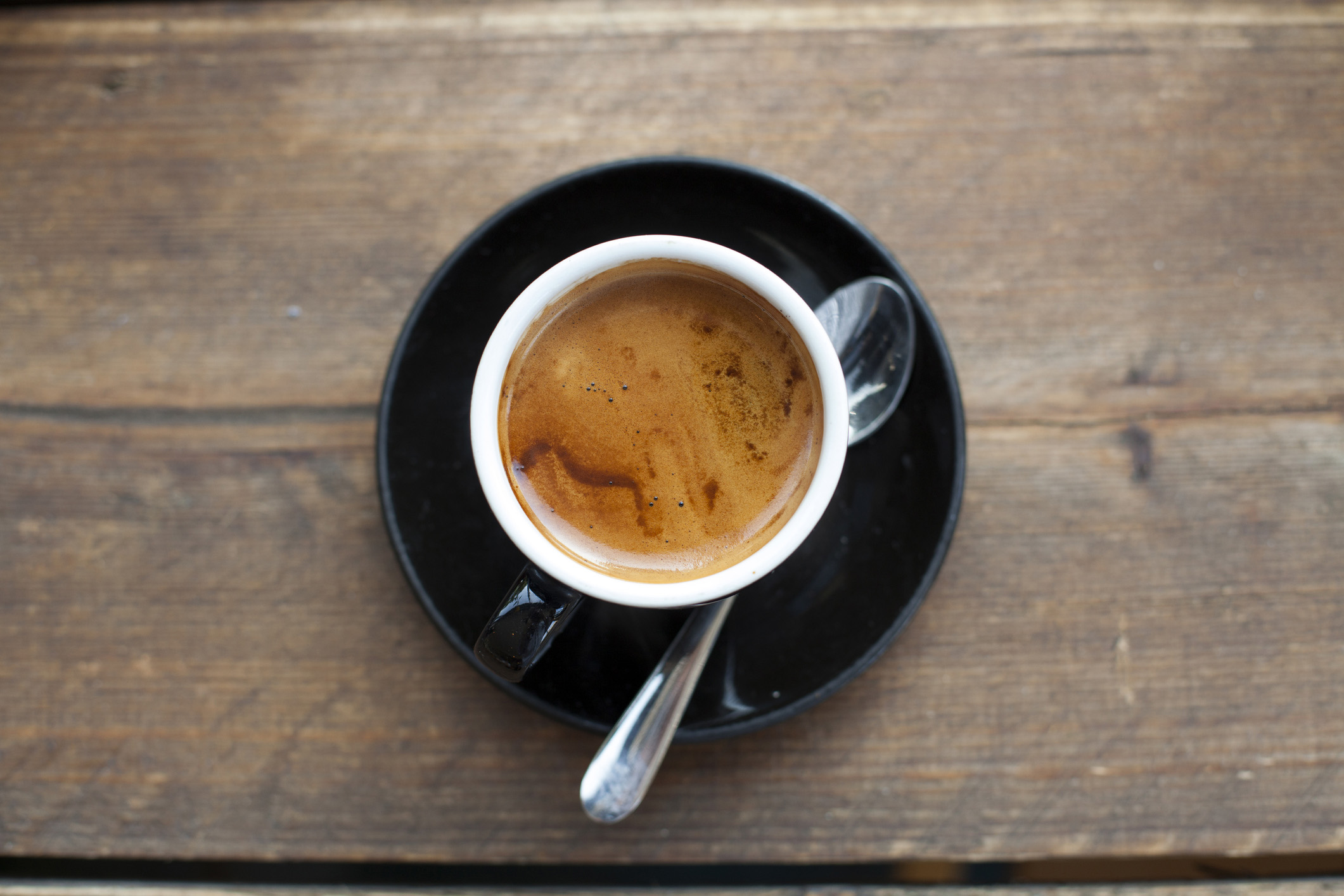 A small cup of espresso sits on a black saucer with a teaspoon on a wooden tabletop